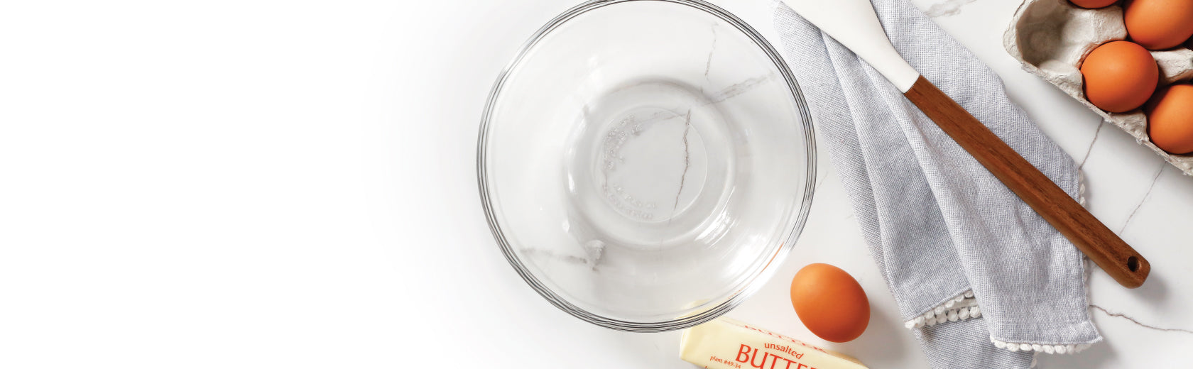 An empty glass bowl on a counter with a spatula, kitchen towel, eggs and butter around the bowl.