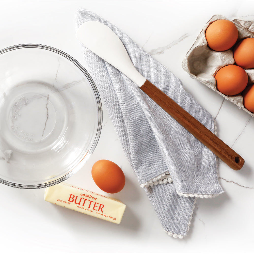 An empty glass bowl on a counter with a spatula, kitchen towel, eggs and butter around the bowl.